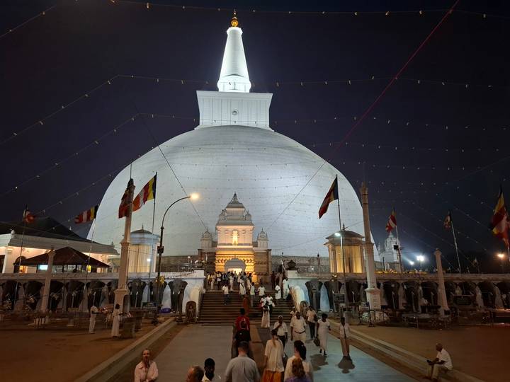 A giant white stupa illuminated at night with worshippers at its base.