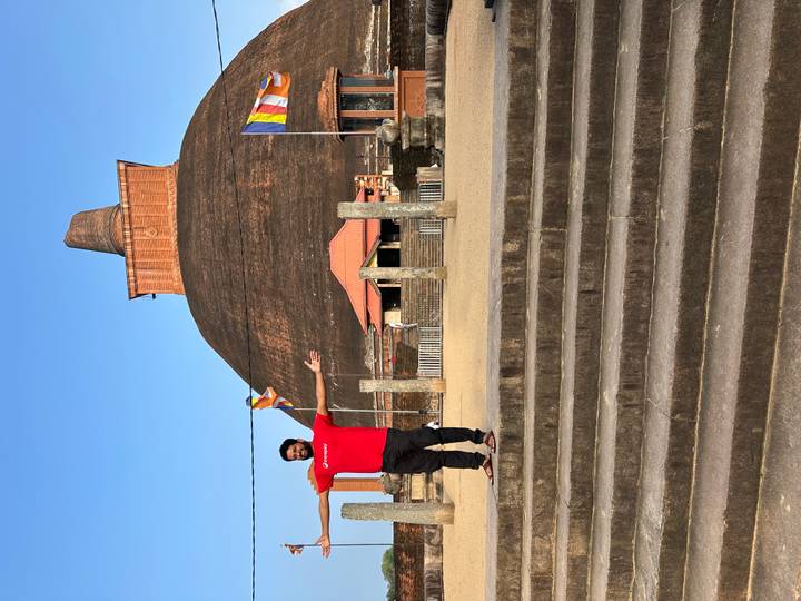 A traveler spreads his arms in front of a massive brick dagoba under blue sky.