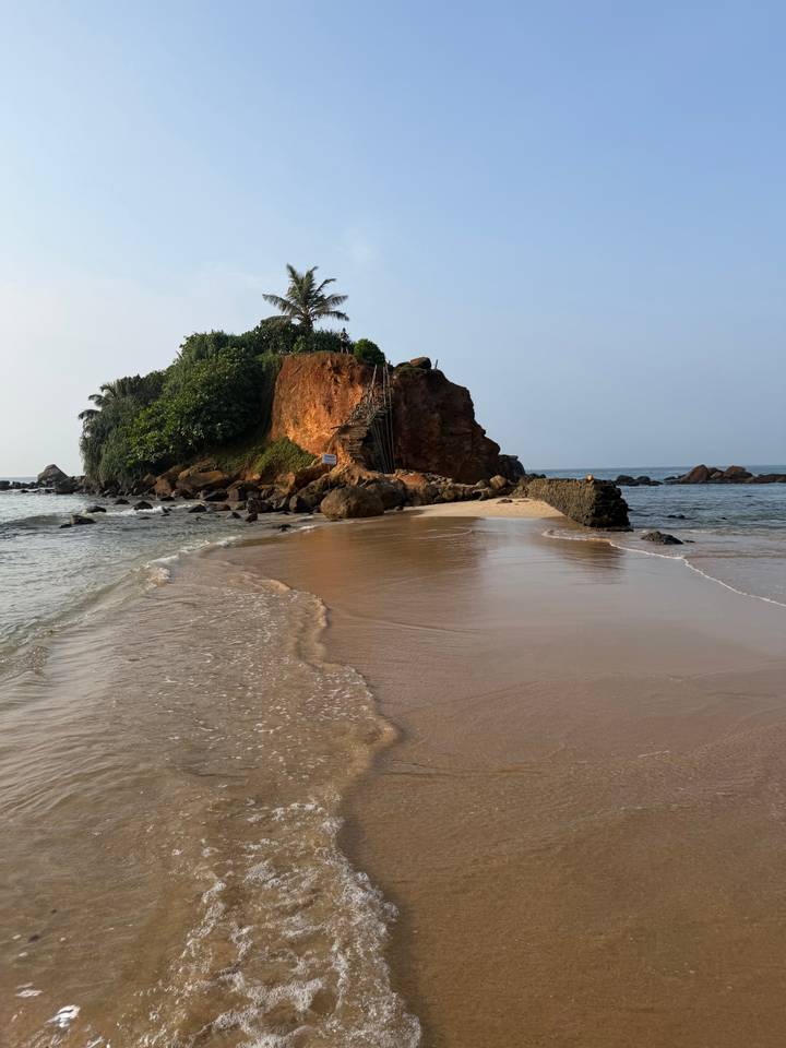 A narrow sand spit leading to a rocky islet topped with greenery.