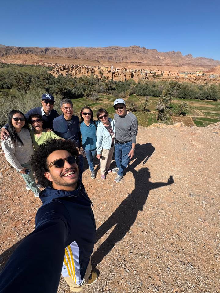 Smiling tour group posing on a rocky viewpoint above a date-palm valley.