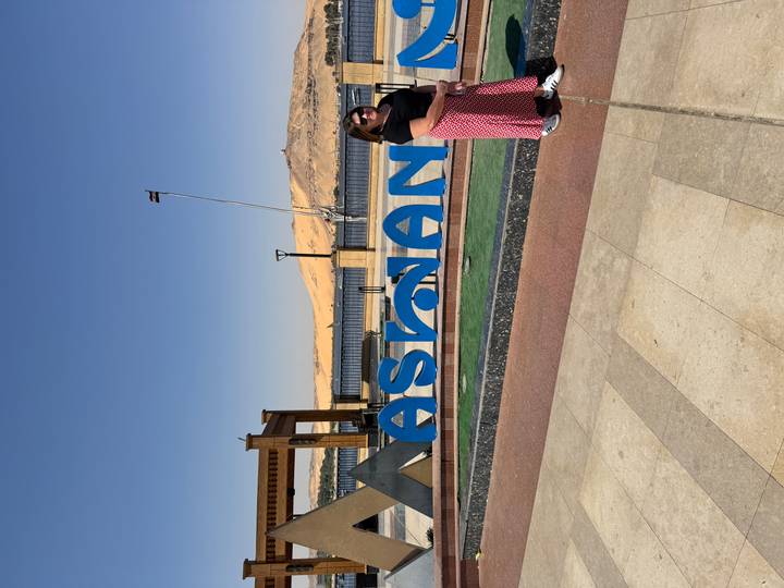 Smiling woman next to a bright blue Aswan sign with river and sand dunes in view.