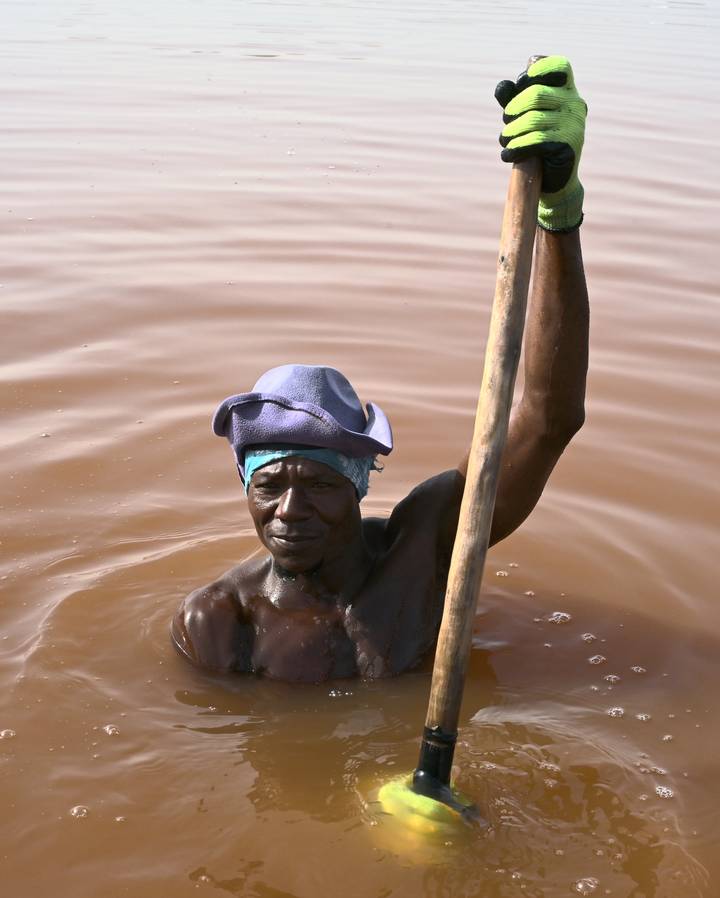 Man chest-deep in milky pink water holding a wooden pole, wearing a scarf and floppy hat.