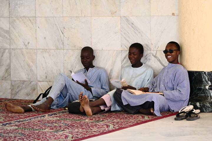 Three teenage boys in loose robes study while sitting barefoot against a marble wall on a patterned carpet.