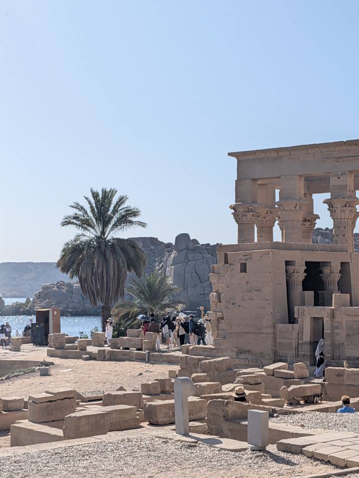 Ancient Egyptian temple columns beside a palm-lined riverside with visitors exploring under bright sky.