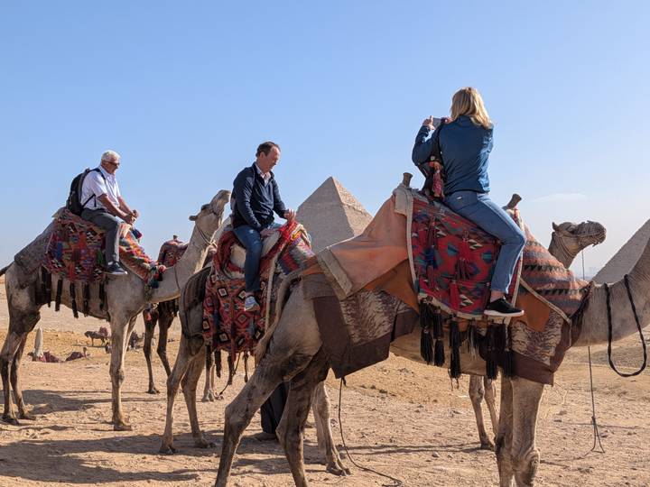 Tourists riding camels with the pyramids rising from the Giza desert in the background.