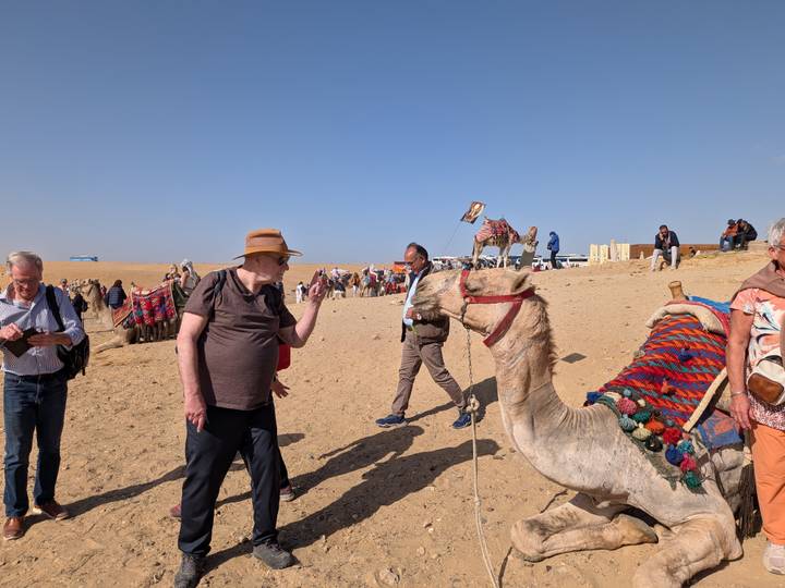 Busy scene of travellers and camel handlers on the sandy desert near the pyramids.