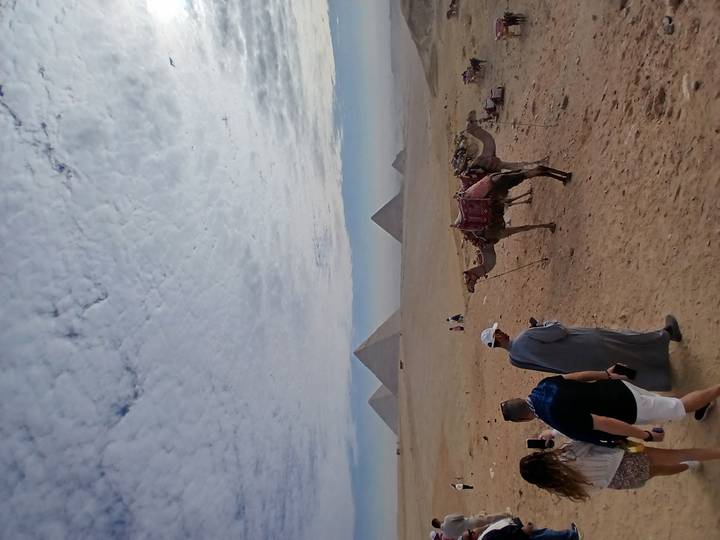 Camels and tourists scattered across a hazy desert foreground with pyramids piercing a dramatic cloud-filled sky.