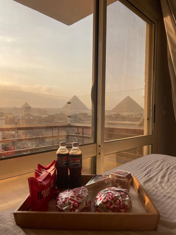 Hotel-room view of the Giza Pyramids at sunset with two chilled Pepsi bottles on the windowsill.