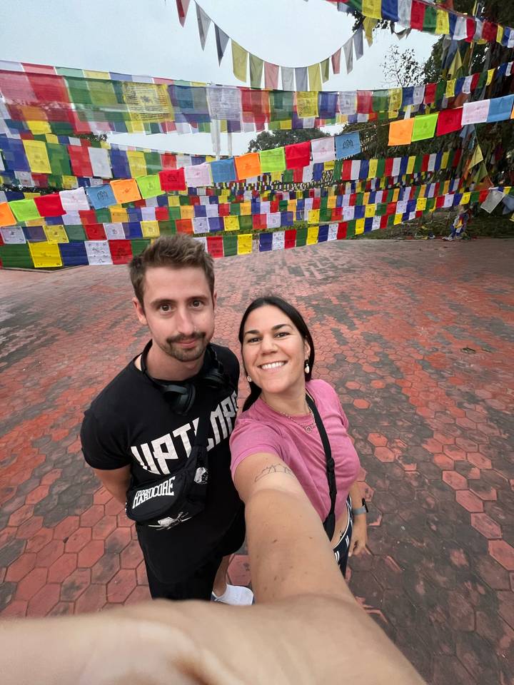 Couple selfie in front of colorful prayer flags on a paved courtyard in Nepal.