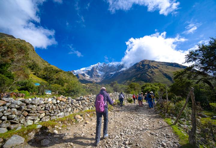 Backpackers hike a rocky trail toward snow-covered Andean peaks under a vivid blue sky.
