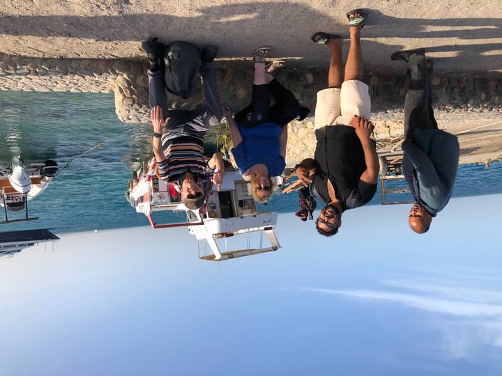 Travelers make peace signs while waiting near small boats on a sunny seaside jetty.