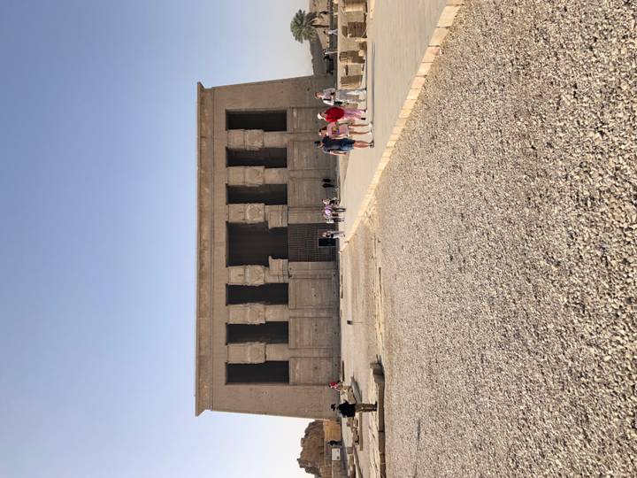 Visitors approach a massive stone facade of an ancient Egyptian temple on a bright sunny day.