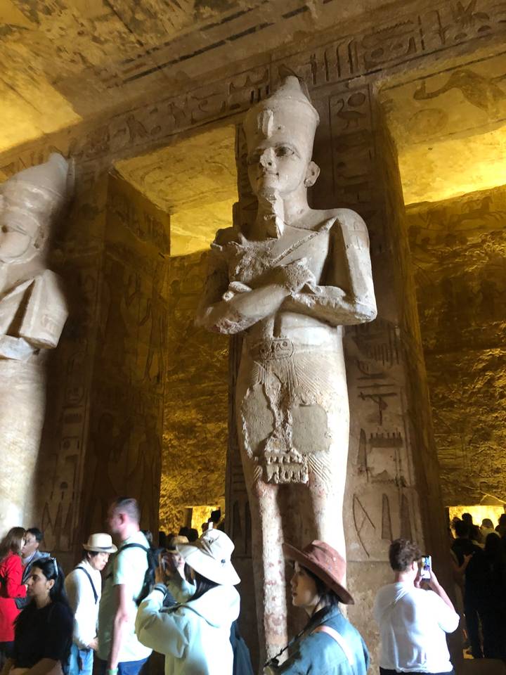 Interior view of a dimly lit temple chamber featuring a large stone statue of a pharaoh with crossed arms.