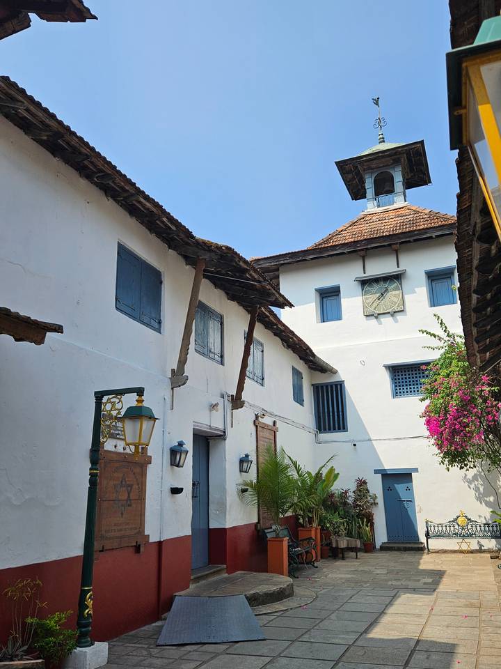 Whitewashed courtyard facade with blue wooden windows and a clock, typical of Kochi’s Paradesi Synagogue, under clear blue sky.