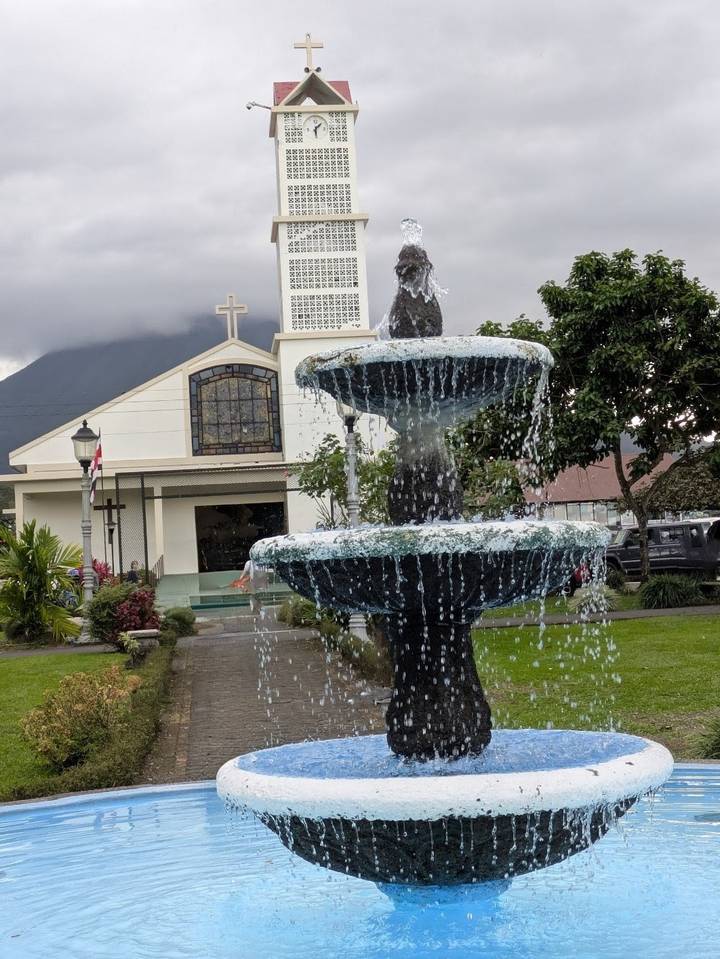 Fountain splashing in front of a church with a cloud-covered volcano in the background.