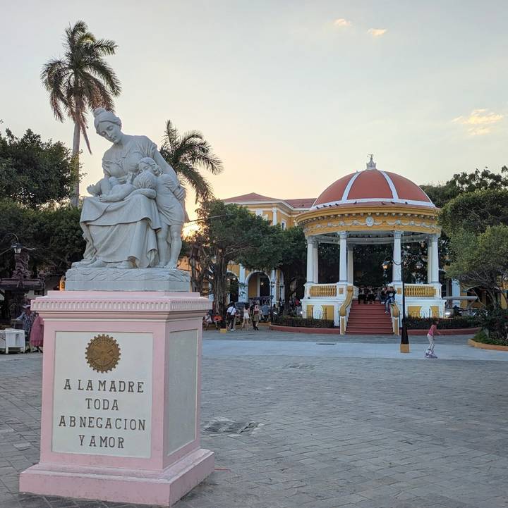 Town plaza at sunset featuring a mother-child statue, a bandstand, and relaxed locals.