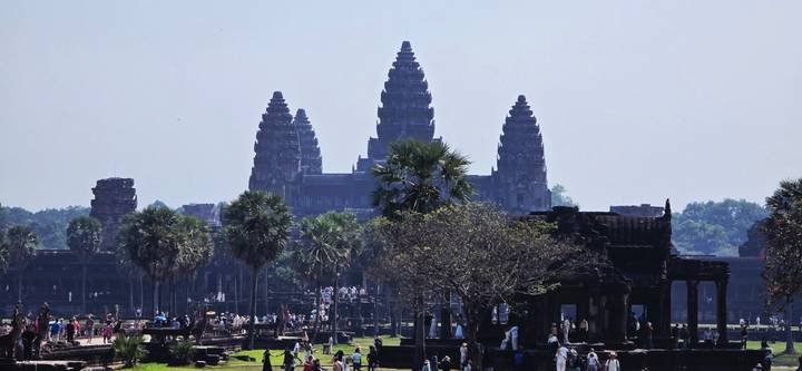 Angkor Wat's iconic towers rise above palm trees and crowds of visitors in clear daylight.