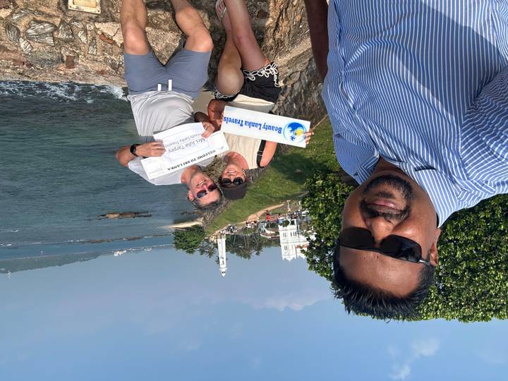 A smiling trio poses for a selfie above a coastal fort and lighthouse under clear blue skies.