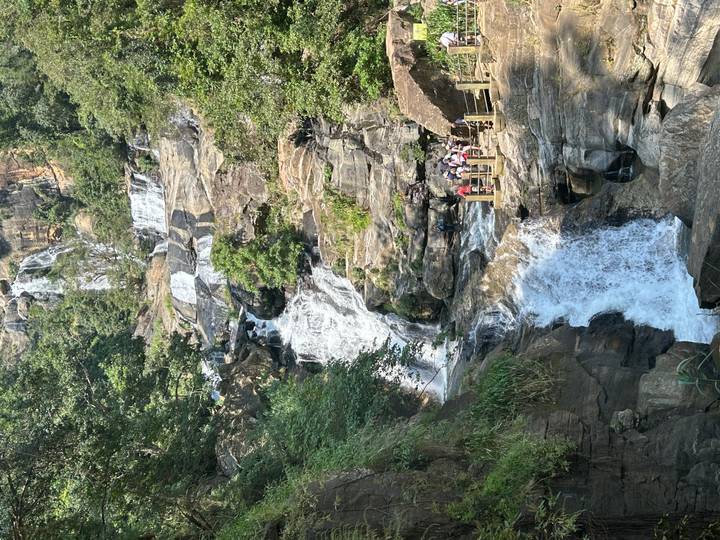 A multi-tiered waterfall tumbles through jungle cliffs as visitors gather on a viewing deck.