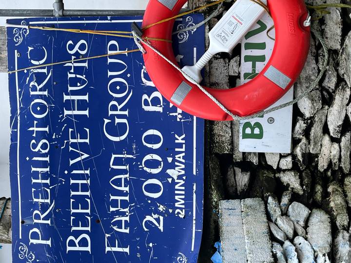 Weather-worn blue information sign for prehistoric Beehive Huts with a red life ring hanging beside it.