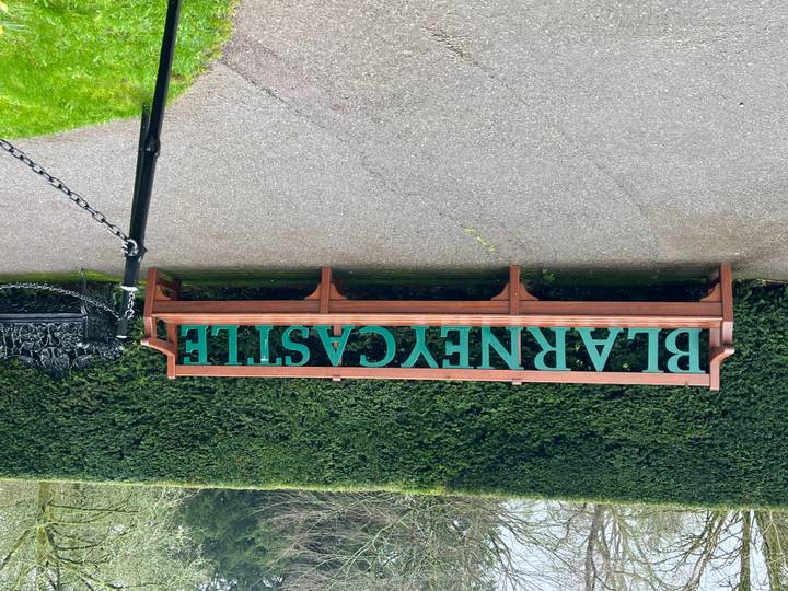 Wooden bench with large green letters spelling 'BLARNEY CASTLE' set against tall manicured hedges.
