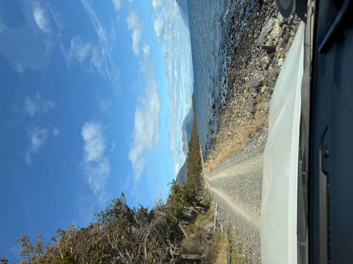 Gravel coastal road beside a blue lake with distant mountains viewed from a vehicle bonnet on a bright day.
