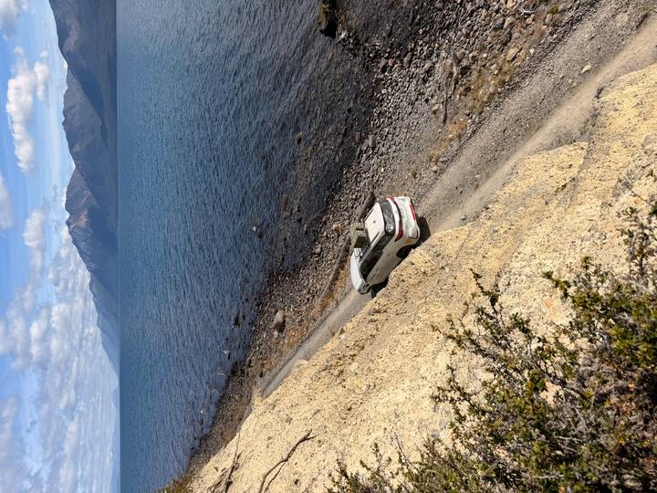 White 4x4 drives a narrow dirt track along a vast lake with snow-capped mountains in background.