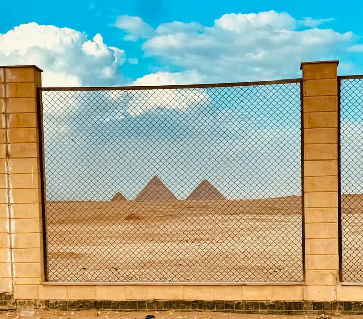 View of three pyramids of Giza distant behind a chain-link fence on a hazy sunny day.