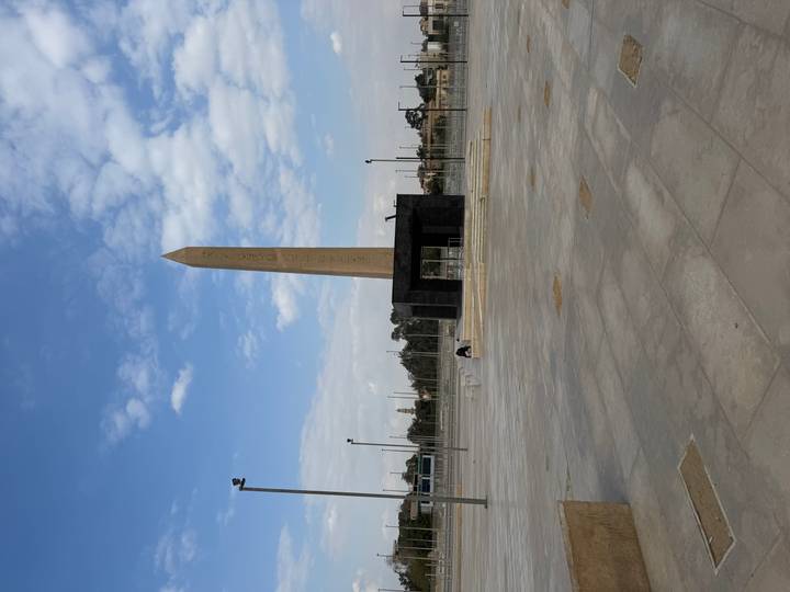 Tall stone obelisk stands in an open modern plaza under partly cloudy sky.