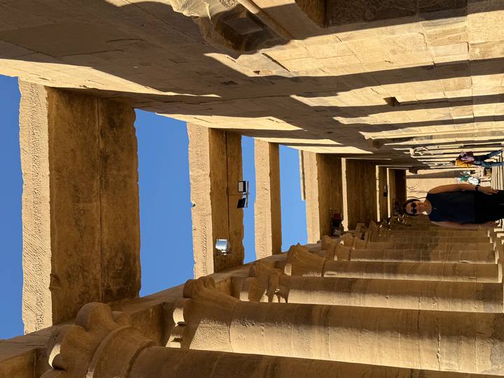Visitor stands beneath stone architraves and column rows in bright sunlight within temple corridor.