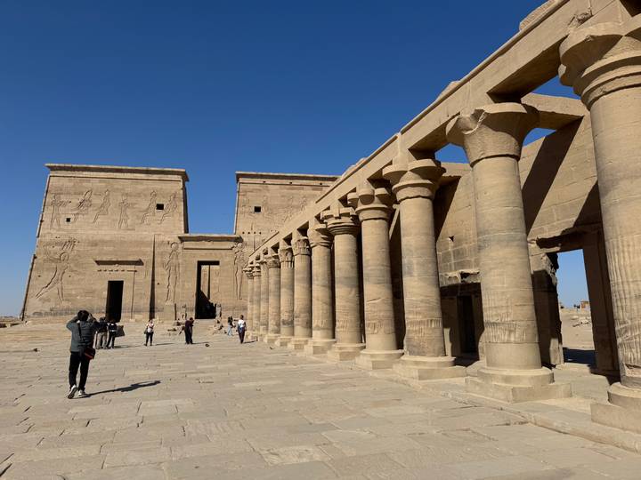 Open stone courtyard with row of tall columns and hieroglyphs, visitors scattered under blue sky.