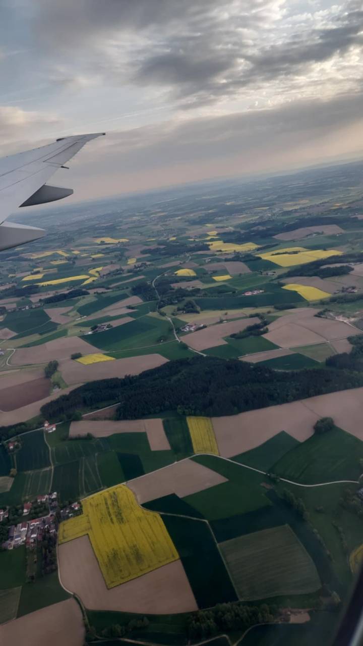 View from airplane window of patchwork farmland fields and forests below a cloudy sky.
