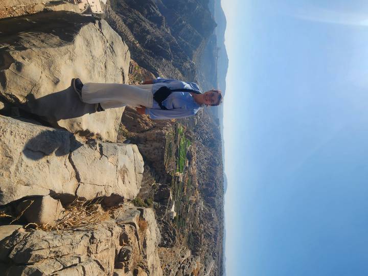 Traveler standing on dramatic cliff edge overlooking deep canyon and mountain plateau under bright blue sky.