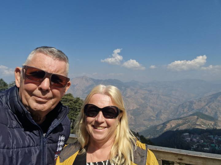 Couple posing at a mountain viewpoint with expansive Himalayan ranges and blue sky behind them.