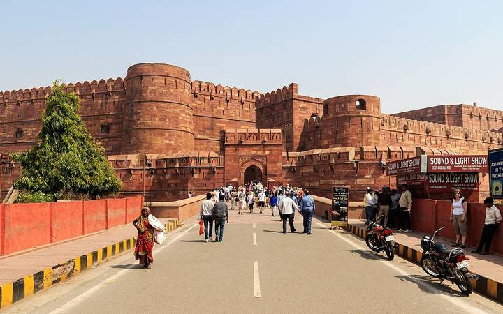 Entrance causeway to massive red-sandstone Agra Fort bustling with tourists on a sunny day.