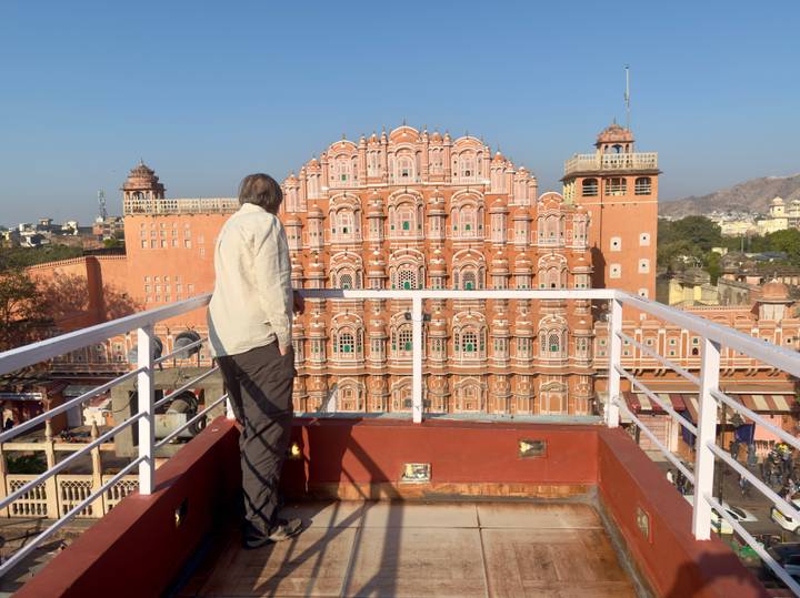 Man on a rooftop terrace gazes at the intricate pink façade of the Hawa Mahal against a clear blue sky.