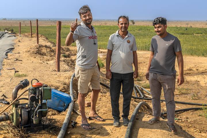 Three men pose cheerfully beside irrigation equipment in a dry rural field.