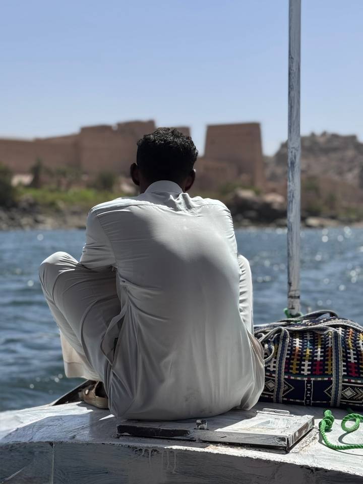 Man in white robe sits on the edge of a felucca drifting along the Nile with temple ruins blurred in the background.