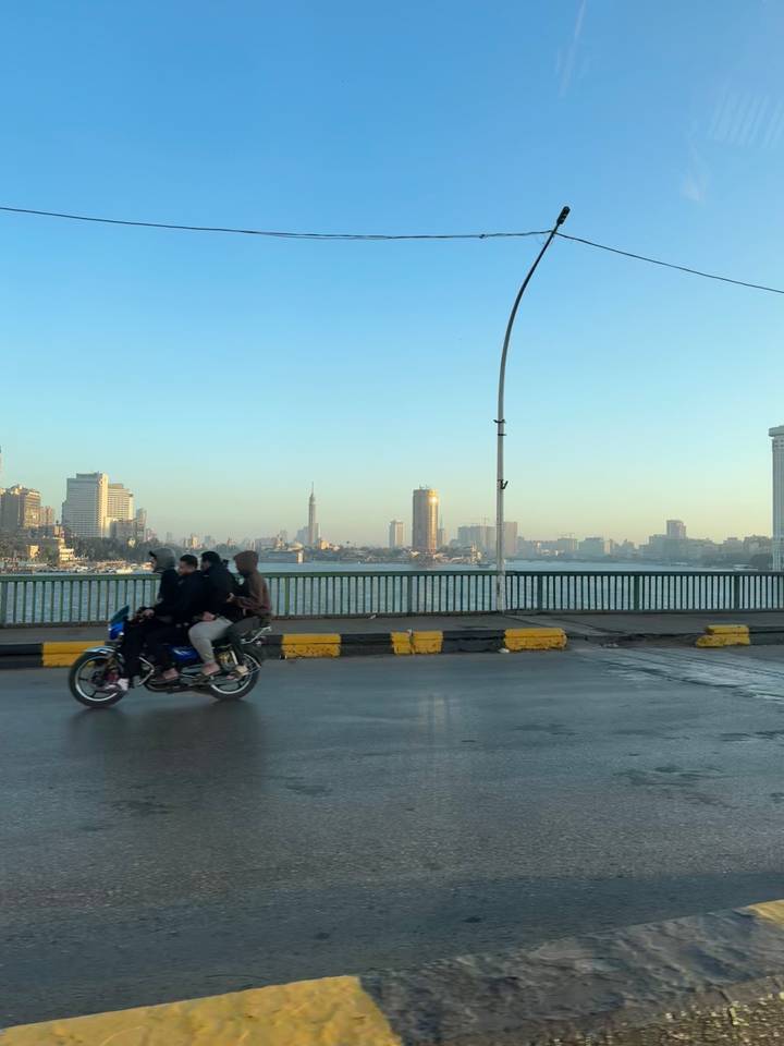 Three riders on a motorcycle cross a bridge over the Nile with Cairo’s skyline in soft morning light.