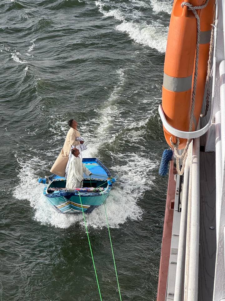 Two vendors in traditional robes stand upright in a small blue boat navigating choppy Nile waters alongside a cruise ship.