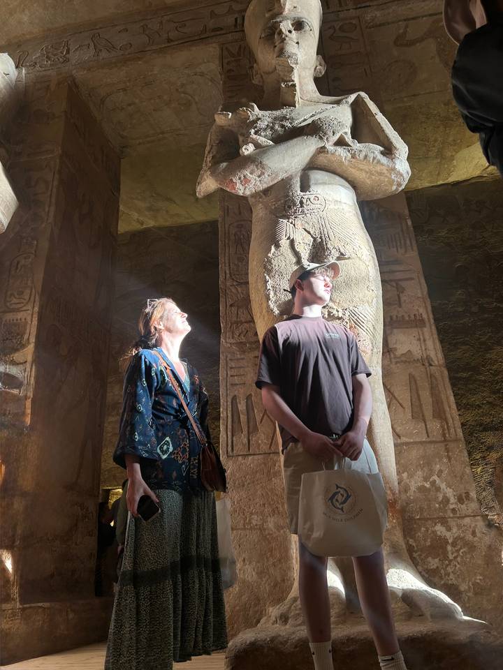 Sunbeams filter through a temple column illuminating two visitors admiring ancient hieroglyphs.