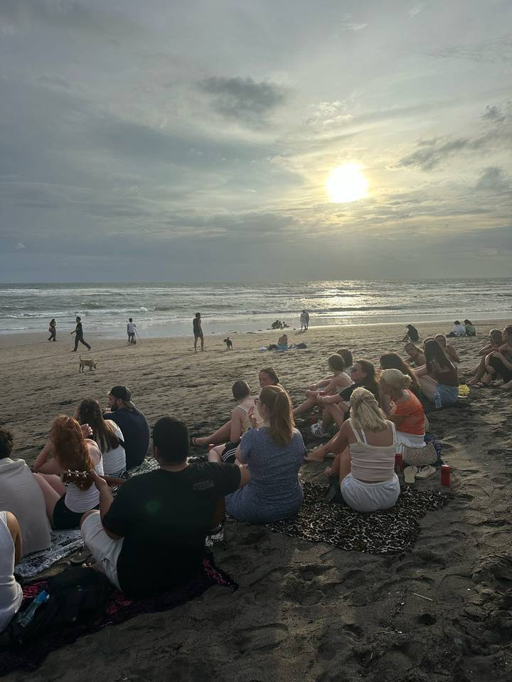 Dozens of travellers sit on a dark sandy beach watching the sun set over rolling surf.