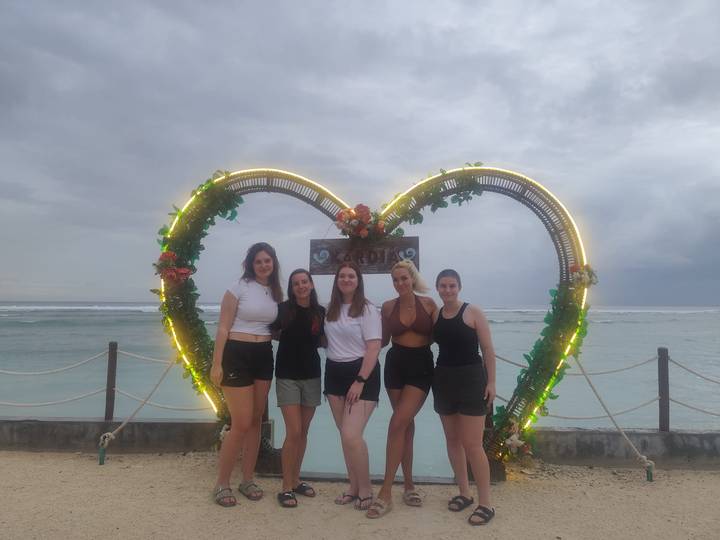 Five friends stand in front of a heart-shaped floral frame lit with lights on a tropical shoreline.