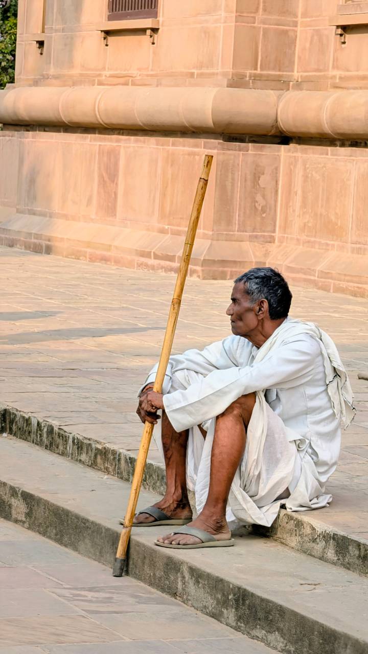 Elderly man in simple white clothing sits on stone steps holding a bamboo walking stick, gazing into the distance.