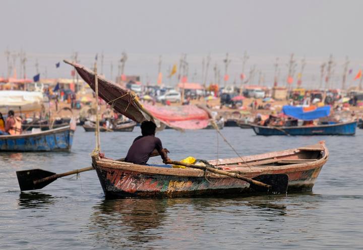 Man rows a weathered wooden boat on a wide river with a busy, colorful festival shoreline in the background.