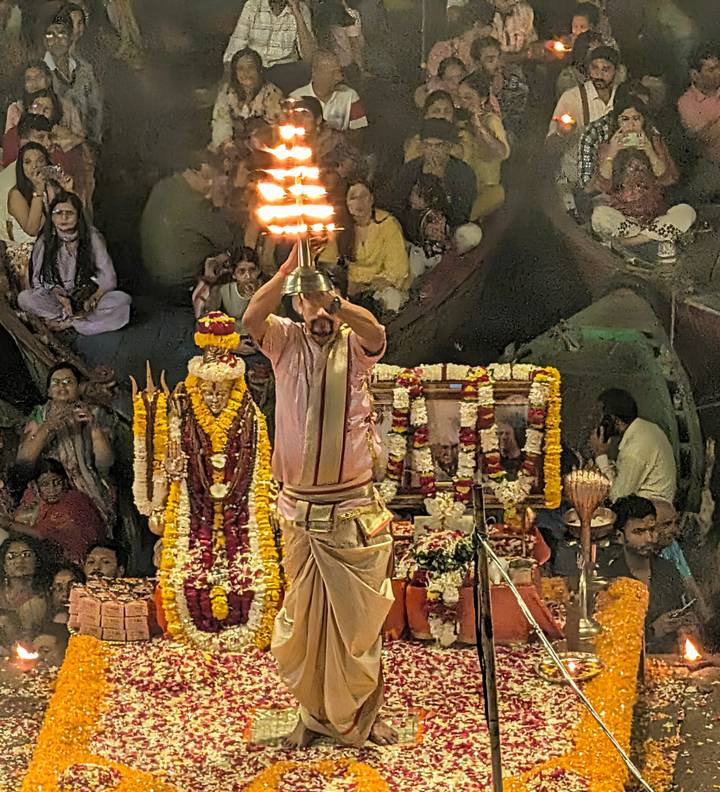 Priest raises a multi-tiered oil lamp during a crowded evening Ganga Aarti ritual surrounded by marigold garlands and worshippers.