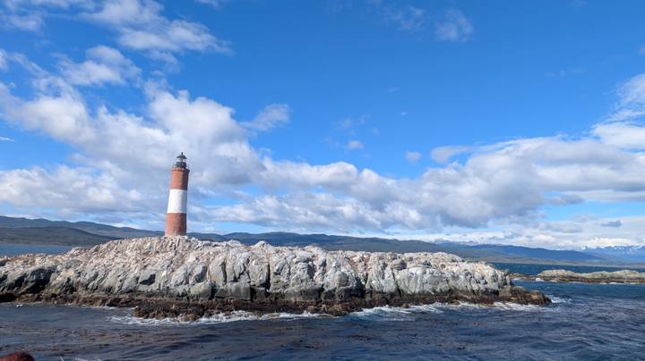 Red-and-white Les Éclaireurs lighthouse stands on a rugged rocky islet under a vivid blue sky with scattered clouds.