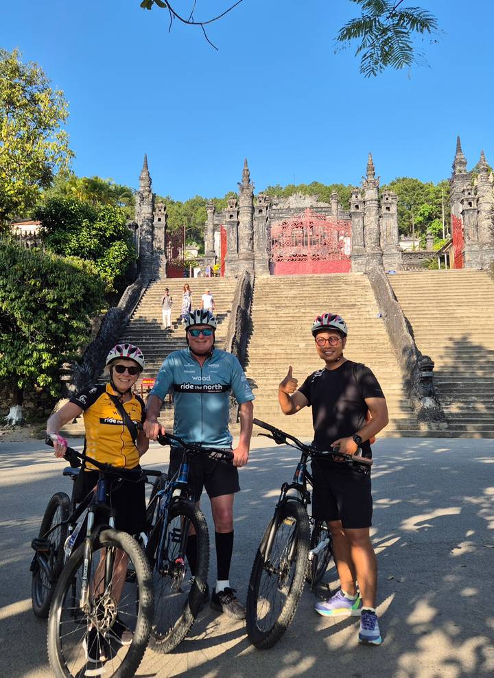 Three cyclists pose proudly with bikes in front of the monumental stairs of Khai Dinh Tomb.