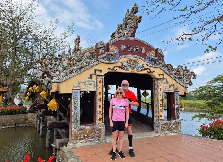 Smiling pair of cyclists pose beside the ornately decorated Thanh Toan covered bridge.