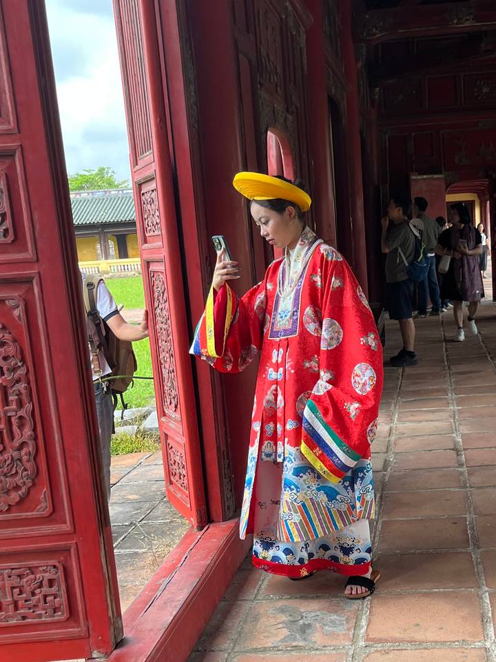 Woman in ornate royal costume takes a selfie inside a richly carved red wooden corridor.